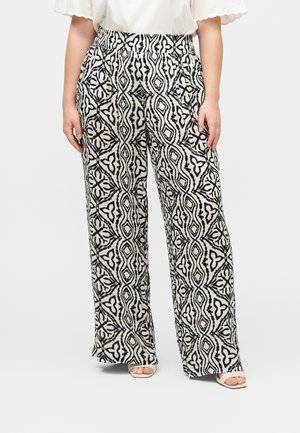 Pantalon à jambes larges avec motif géométrique noir et blanc porté avec un haut blanc et des sandales ouvertes, mains reposant sur les côtés.