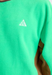 Zeleni flis pulover s trikotnim belim logotipom in rjavimi akcenti na rokavih. Mehka tekstura, zmerna debelina in sproščen kroj.