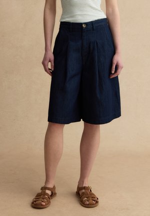 Persoon die donkerblauwe knielange geplooide shorts, een wit gestreept mouwloos topje en bruine sandalen met gesloten tenen draagt, staand op een beige achtergrond.