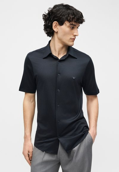 Άνδρας με σγουρά μαλλιά που φοράει μαύρο πουκάμισο με κοντά μανίκια και γκρι παντελόνι, κοιτάζει προς τα δεξιά του έχοντας ένα χέρι στην τσέπη του.