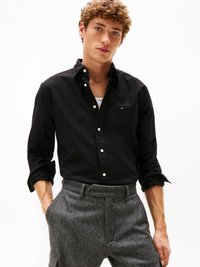 Camisa preta de botões com colarinho, apresentando um pequeno logo no peito. Combinada com calças em espinha de peixe cinzas. Textura suave.