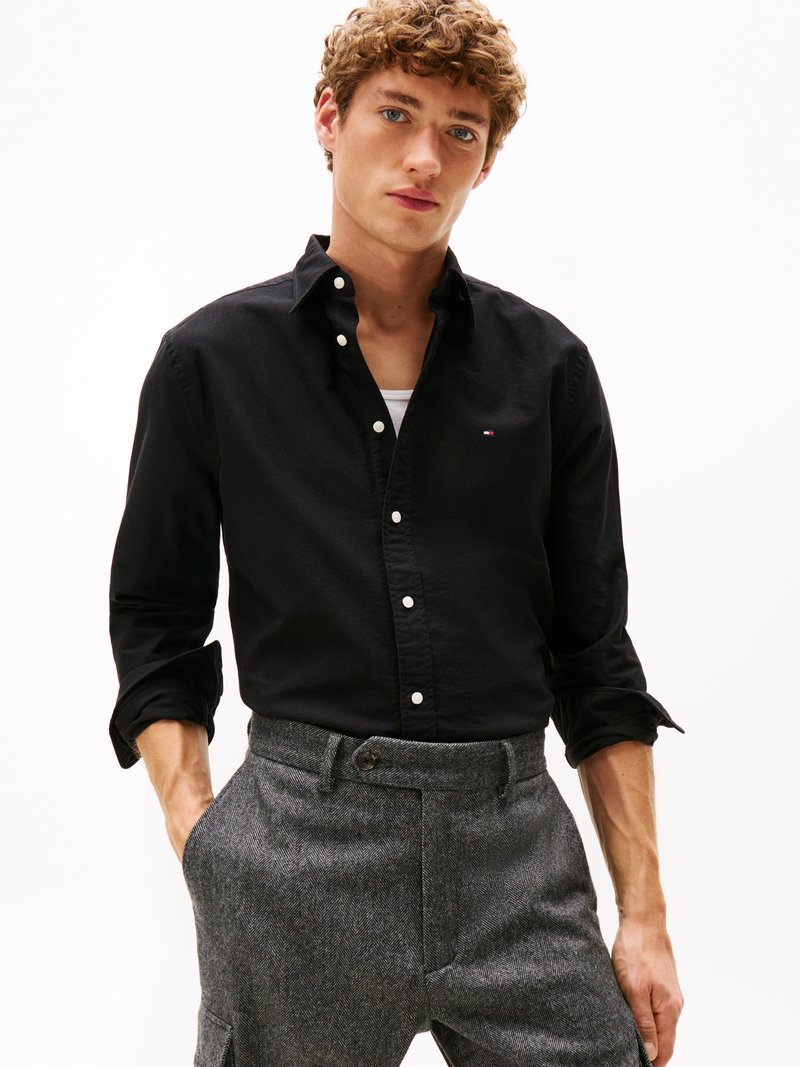 Camisa preta de botões com colarinho, apresentando um pequeno logo no peito. Combinada com calças em espinha de peixe cinzas. Textura suave.