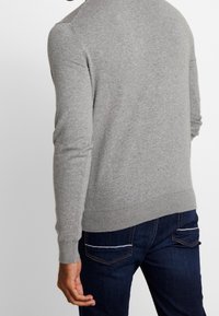 Grå stickad tröja med långa ärmar, som har en rundad halsringning och ribbade muddar. Matchad med mörka jeans, som visar bakifrån.