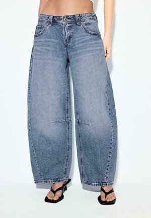 Široké vyblednuté modré rifle z denimu nosené s čiernymi sandálmi na podpätku so štvorcovou špičkou, skrátené v páse, odhaľujúce holý pás.