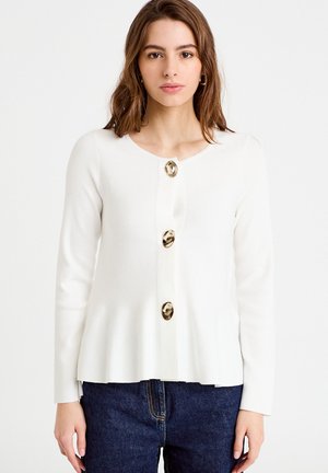 Donna con un cardigan bianco a maniche lunghe con tre grandi bottoni dorati, abbinato a jeans di denim blu scuro, in piedi davanti a uno sfondo semplice.