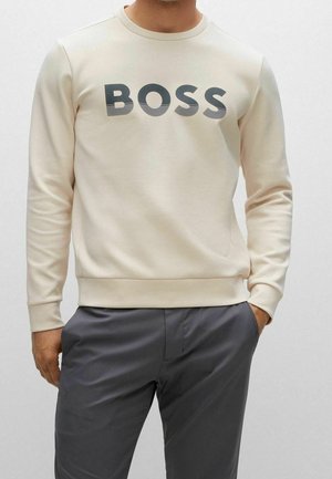 Mann trägt einen beigen Sweatshirt mit der Aufschrift "BOSS" in Schwarz und Grau, kombiniert mit dunkelgrauen Hosen, eine Hand in der Tasche, vor einfachem Hintergrund.