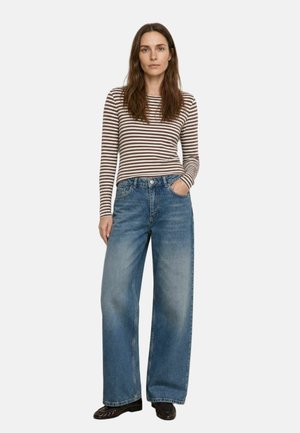 Donna con camicia a righe marroni e bianche a maniche lunghe e jeans blu a gamba larga, in piedi con una mano in tasca su sfondo neutro.