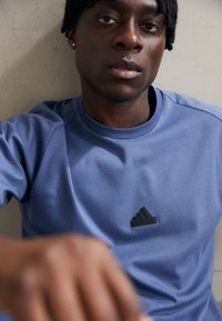 Niebieska bawełniana koszulka z ribbowym dekoltem, z czarnym logo Adidas na klatce piersiowej, gładką strukturą i krótkimi rękawami raglanowymi.