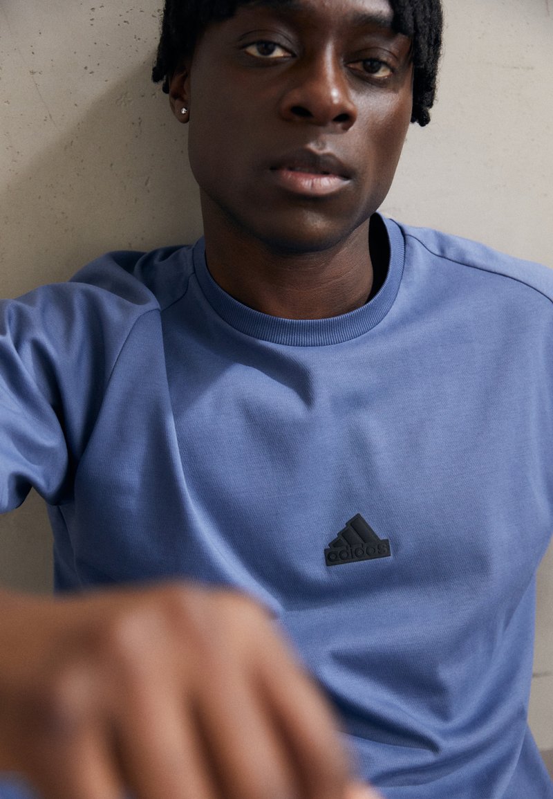 Niebieska bawełniana koszulka z ribbowym dekoltem, z czarnym logo Adidas na klatce piersiowej, gładką strukturą i krótkimi rękawami raglanowymi.