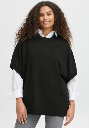 Maglione nero oversize con maniche corte, orlo a costine e dettagli con bottoni laterali. Indossato sopra una camicia bianca con colletto e abbinato a jeans grigi.