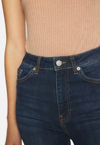 Primer plano de una persona que lleva jeans de mezclilla azul oscuro con costuras naranjas y una camiseta sin mangas de canalé beige metida por dentro.