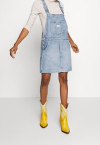 Denimklänning i ljusblått, med justerbara axelremmar, framficka och sidoknappar. Matchas med klara gula cowboystövlar.