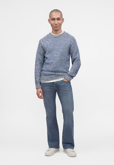 Homme portant un pull en laine bleu, un jean bleu clair et des baskets blanches, debout avec une main dans la poche devant un fond blanc uni.