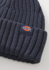 Sötét tengerészkék kötött sapka bordás textúrával és hajtott mandzsettával, piros, sárga és fehér kör alakú Dickies logóval.
