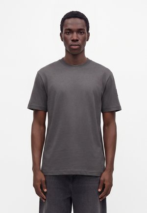 Ung mand med kort krøllet hår står med fronten mod kamera iført en ensfarvet mørkegrå kortærmet t-shirt og mørke bukser mod en hvid baggrund.