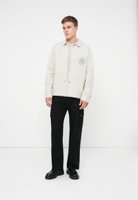 Giacca beige chiaro con bottoni a pressione e logo sul petto, abbinata a pantaloni cargo neri. L'outfit presenta un aspetto stratificato e texturizzato.
