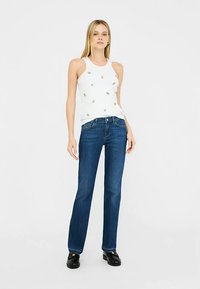 Top bianco senza maniche con piccoli ornamenti, abbinato a jeans blu scuro a gamba dritta. Modella in piedi con le mani sui fianchi, scarpe nere visibili.