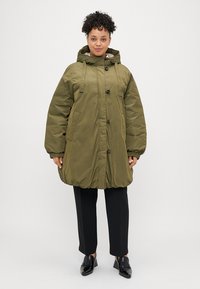 Alyvuogių spalvos, per didelis pūkinis paltas su kapišonu, priekiniais mygtukais, šoninėmis kišenėmis ir reguliuojamomis raištelėmis. Lygi tekstūra ir lengvas dizainas.