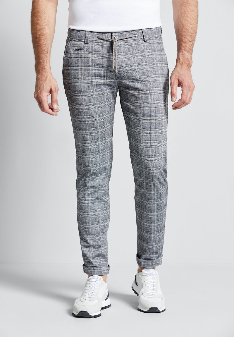 Homme portant un pantalon gris à carreaux coupe slim avec ourlets retroussés et des baskets blanches, se tenant devant un fond clair uni.