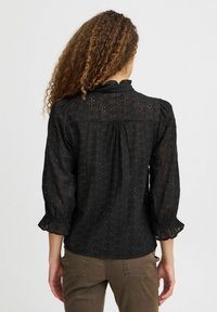 Blusa nera con un design testurizzato e ricamato, maniche 3/4, polsini arricciati e scollo alto. Presenta un dettaglio con pieghe sul retro.