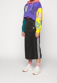 Sweatshirt cropped colorato con grafiche, gonna midi nera con strisce bianche laterali e sneakers chiare con accentuazioni testurizzate.