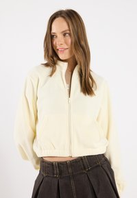 Cropped fleece jacket in soft yellow, featuring a full zip, stand collar, and front pockets. Paired with a dark pleated skirt.