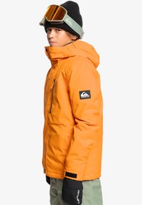 Veste isolante orange avec capuche, poche zippée sur le côté et patch logo noir. Associée à des gants noirs et à des lunettes de protection, portée par une jeune personne.