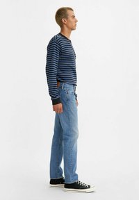 Camisa de manga larga a rayas en azul marino y azul, combinada con jeans rectos de un azul claro y zapatillas altas de color negro. Vista de perfil lateral.