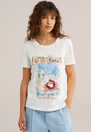 Lokkis juustega naine, kes kannab valget T-särki, millel on rannamaastik, punane krabi ja tekst "L'île de Beauté", ning helerohelisi pükse neutraalse taustaga.