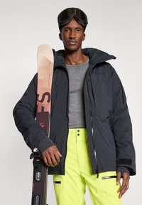 Mies, joka on pukeutunut mustaan hiihtotakkiin, neonkeltaisiin housuihin ja suojalaseihin, pitää pinkkejä ja mustia suksia pystyasennossa valkoista taustaa vasten.