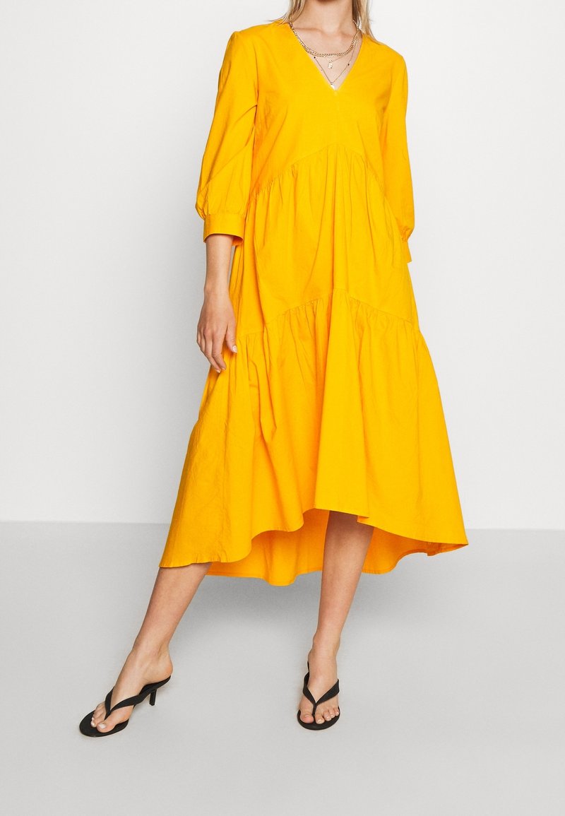 Femme portant une robe midi jaune vif avec un col en V, des manches trois-quarts et des sandales à talons noires, sur fond blanc.