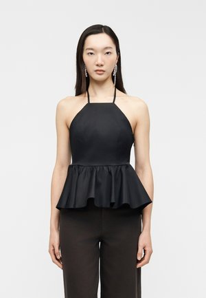 Kvinde med langt mørkt hår iført en ærmeløs sort haltertop med peplumkant og hængende krystaleøreringe, stående mod en ensfarvet baggrund.