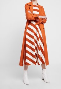 Un trench-coat orange sur une robe blanche avec des rayures horizontales en rouille. Des bottines blanches complètent la tenue.