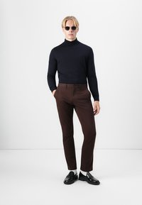 Fekete garbó, barna nadrág, fekete cipő és napszemüveg. A modell egy sima fehér háttér előtt áll.
