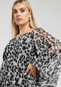 Genomskinlig, oversized topp med svart och grå leopardsnitt, vida ärmar och båtformad halsringning. Texturerad tyg ger visuellt intresse.