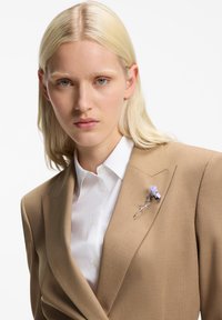 Μπεζ σακάκι πάνω από ένα λευκό πουκάμισο, με ασημένιο φλοράλ καρφίτσα που έχει σχέδιο φωτεινού μωβ λουλουδιού. Ομαλή υφή, προσαρμοσμένη εφαρμογή.