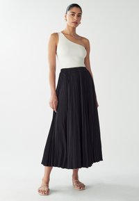 White ribbed one-shoulder top paired with a long black pleated skirt. Model wears simple strappy sandals. Clean, minimal design.
