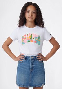 Joven con cabello rizado que lleva una camiseta blanca de Champion con un logo colorido y una falda vaquera azul, con las manos en la cadera.