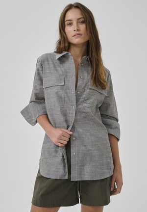 Frau mit langem braunem Haar, die ein grau-weiß gestreiftes Hemd mit hochgekrempelten Ärmeln und dunkelgrüne Shorts trägt.