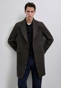 Abrigo de lana marrón oscuro con cuello con muesca, dos bolsillos frontales y botones. Lleva puesto una camiseta polo negra, exhibido sobre un fondo neutral.