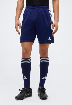 Indossatore maschio in piedi, vestito con pantaloncini da calcio Adidas blu navy, calzettoni abbinati a righe fino al ginocchio e scarpini neri su uno sfondo semplice.