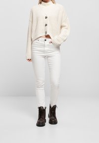 Kvinna i vita högmidjade skinny jeans, kort creamfärgad stickad cardigan med knappar, vit polotröja och mörkbruna snörstövlar.