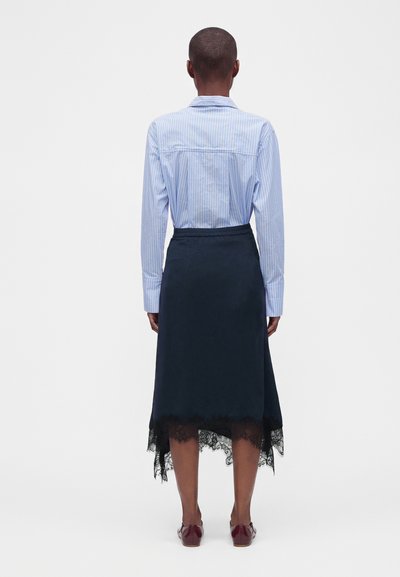 Personne debout de dos, portant une chemise rayée bleue rentrée dans une jupe foncée avec un galon en dentelle et des chaussures plates rouges.