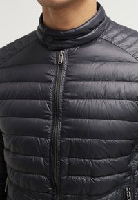 Veste matelassée en doudoune bleu marine foncé. Elle présente un col montant et une fermeture zippée frontale avec des éléments métalliques argentés. Texture lisse et légère.