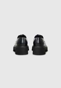Chaussures en cuir noir avec un bout arrondi, semelle en caoutchouc noir épaisse et design minimaliste. L'arrière présente une texture lisse et brillante.