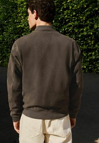Chaqueta bomber de ante marrón con cuello redondo, puños acanalados y cintura ajustada. Presentada en un modelo de espaldas, combinada con pantalones de color claro.