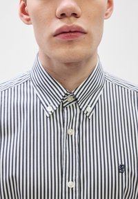 Ριγέ πουκάμισο με κουμπιά σε ναυτικό μπλε και λευκό, με γιακά τύπου spread και λογότυπο στο στήθος. Ομαλό ύφασμα με έντονες κάθετες ρίγες.