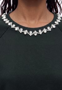 Encolure de vêtement vert foncé ornée de gros strass clairs dans un motif répétitif le long du col.