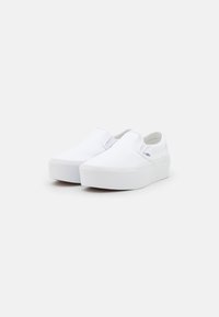 Zapatos slip-on de lona blanca con suela de goma plana, que cuentan con laterales elásticos y un mínimo de marca en el talón. Textura suave y diseño simple.