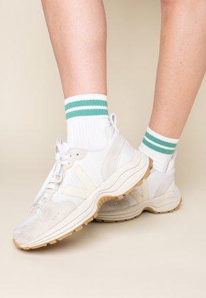 White mesh and suede sneakers with beige accents and rubber sole. Paired with white socks featuring green and white stripes at the top.